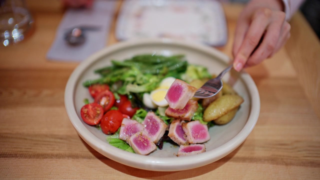 A bowl of salad placed on a wooden table, containing leafy greens, cherry tomatoes, boiled egg slices, and pieces of seared tuna