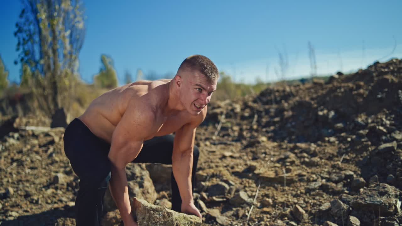 Bodybuilder is training outdoors. Strong athlete with muscular body rising heavy stone and throwing it over his head on the rocky background. Slow motion.
