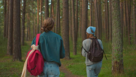 cousins walk along a scenic forest trail filled with lush greenery, one cousin playfully swings her black bag over her shoulder while the other, dressed in a green shirt, watches from behind