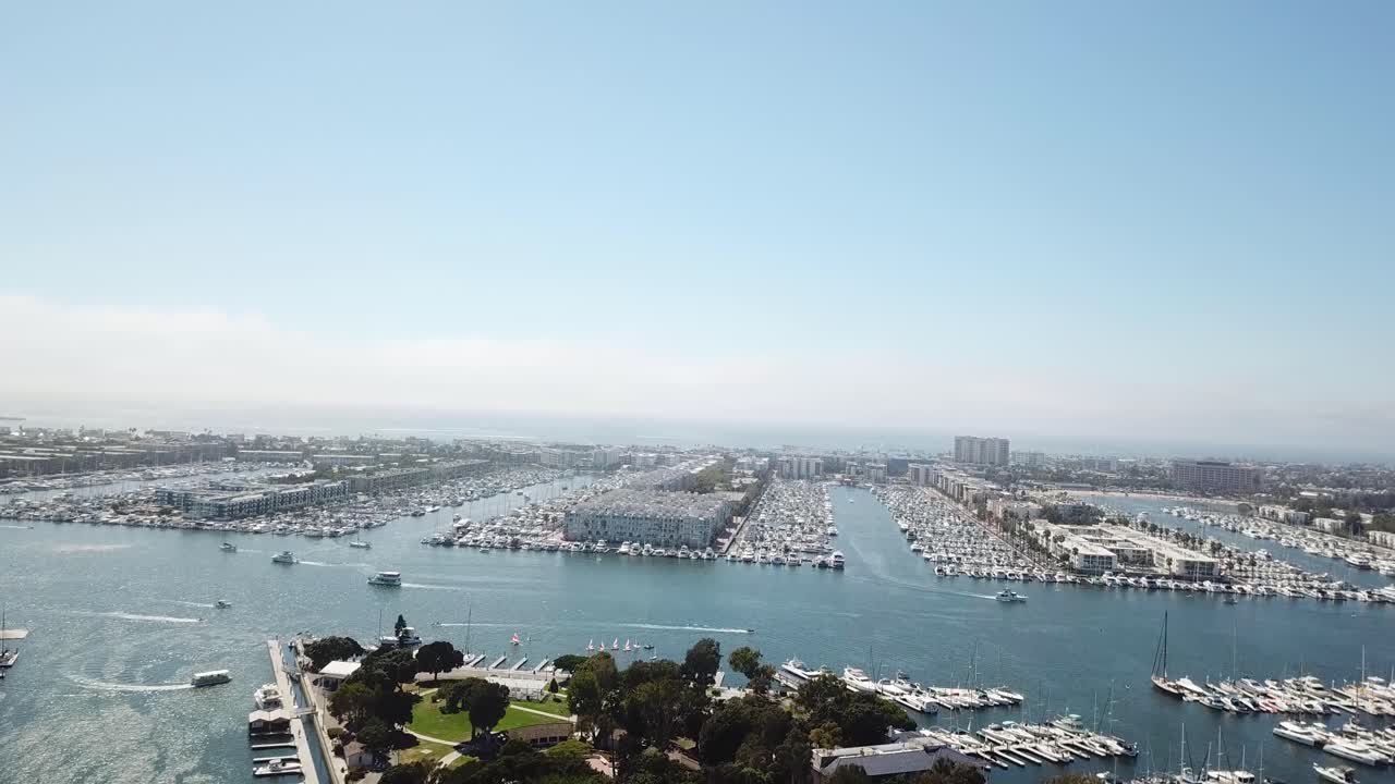 Wide aerial of Marina del Rey, CA. Camera pushes in and ascends, revealing long marina basins packed with yachts, a central navigation channel, and waterfront development under clear SoCal skies