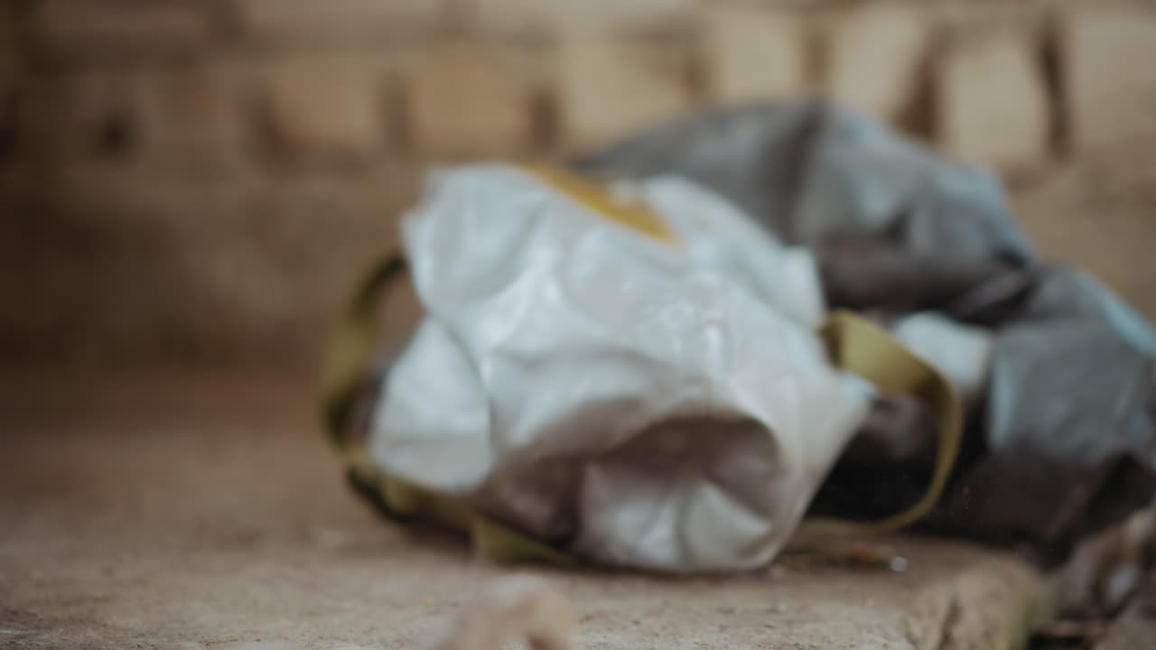 Close up of person dropping protective hazmat mask onto rough dusty ground with worn cracked concrete in muted tones, evoking mood of abandonment