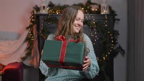 Eine-Frau-Fängt-Und-Schüttelt-Aufgeregt-Ein-Weihnachtsgeschenk,-Das-Mit-Lächeln-Und-Feiertagsdekorationen-Im-Hintergrund-Zu-Hause-Gefüllt-Ist