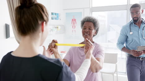 Mujer-Mayor-Diversa-Haciendo-Ejercicio-Con-Banda-Y-Doctora-Asesorando-En-El-Hospital,-Cámara-Lenta