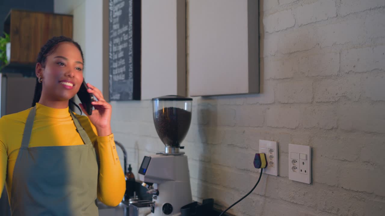 Afroamerikansk barista med forklæde tager telefonen og taster detaljer ind på sin tablet ved cafédisken