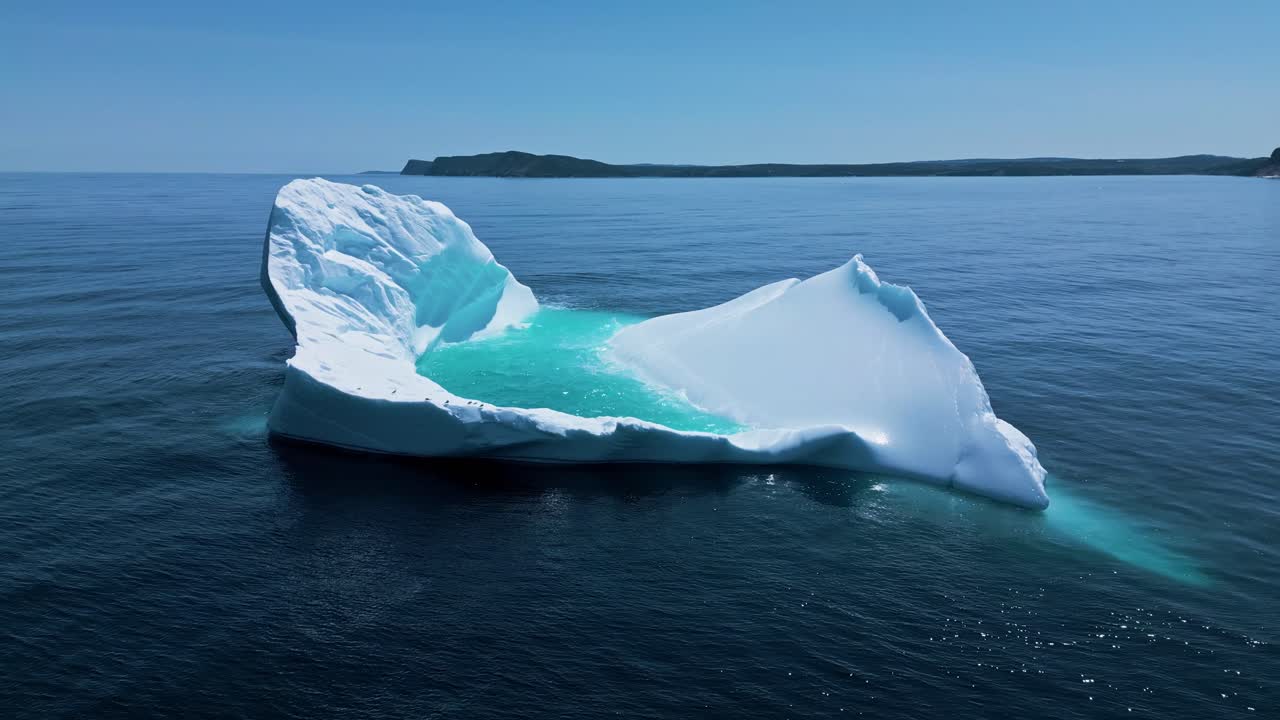 Aerial footage of a large iceberg off Flatrock, Newfoundland, featuring vivid turquoise meltwater in calm Atlantic waters. Birds rest on the iceberg, getting ready to continue the hunt.