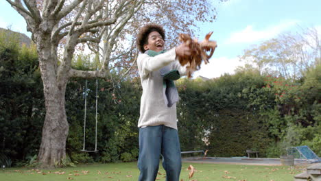 happy african american boy in scarf playing with fallen leaves in garden, slow motion