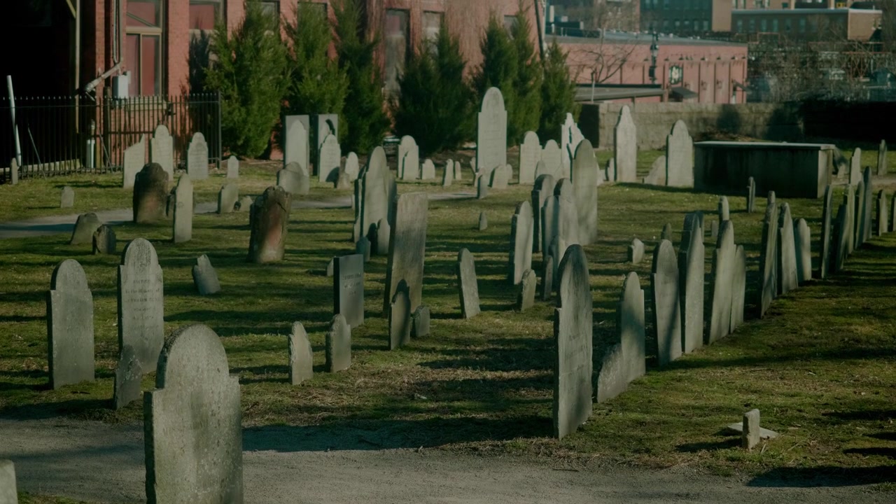 Inquadratura statica media di un cimitero storico a Salem, Massachusetts, con lapidi consumate e lunghe ombre.
