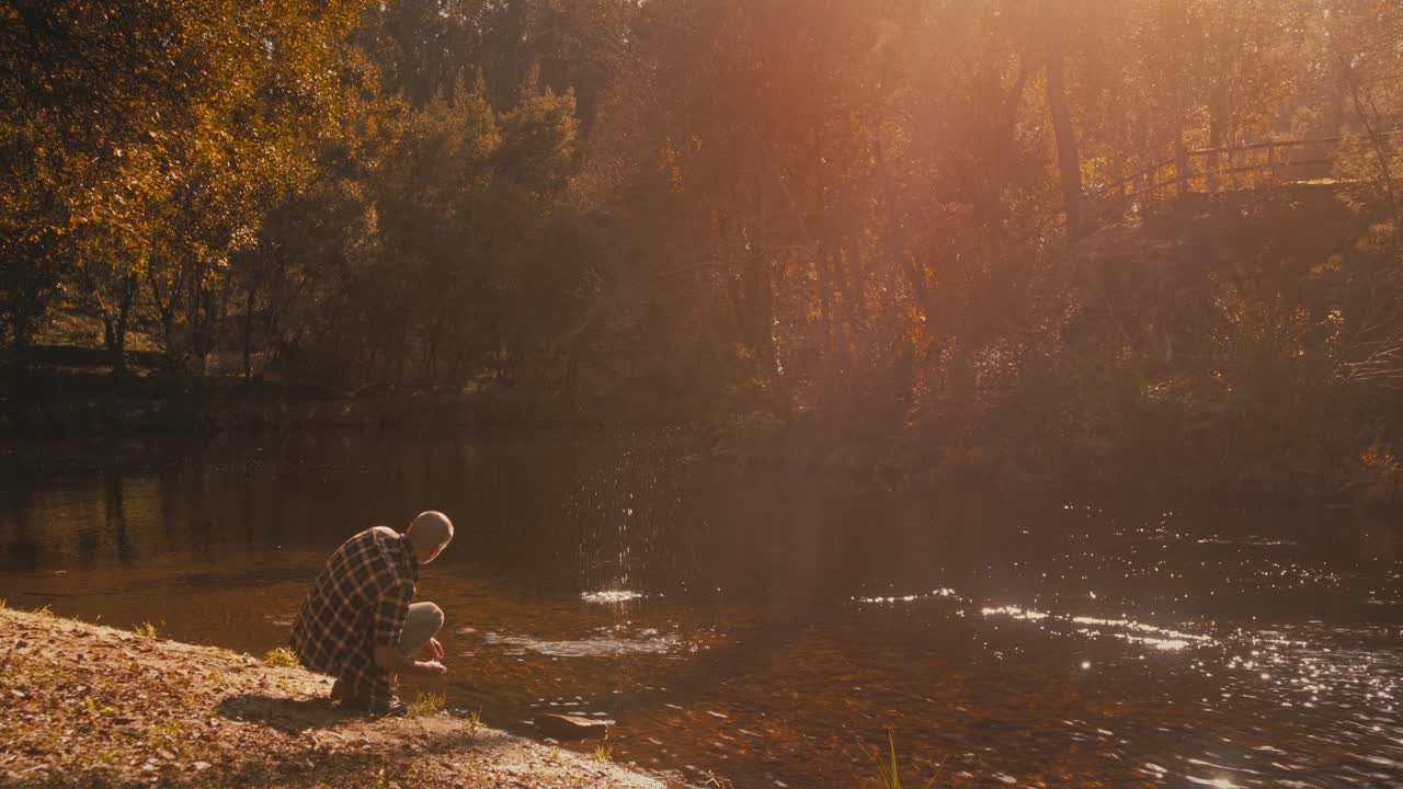 Man meditates by river on sunny day, tossing stones in water. He enjoys nature and solitude. Tranquil clip for travel or self-development.