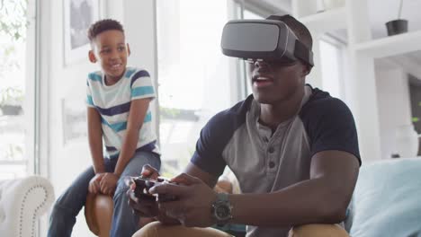 Video-Eines-Glücklichen-Afroamerikanischen-Vaters-Und-Sohnes,-Der-Auf-Dem-Sofa-Sitzt-Und-Mit-Dem-VR-Headset-Spiele-Spielt