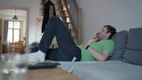 skinny young man sitting on the couch in relaxed position watching tv and looking at his watch to find out what time it is