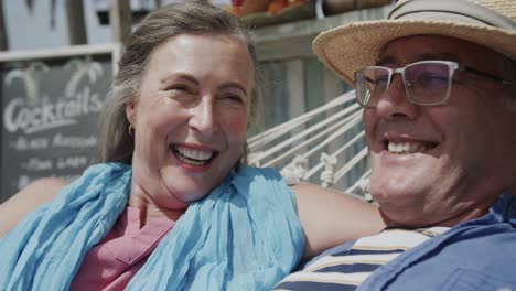 Happy-senior-caucasian-couple-lying-on-hammock-talking-outside-beach-bar,-in-slow-motion