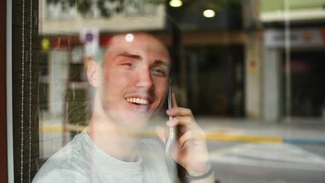 Un-Joven-Alegre-Hablando-Por-Teléfono-Inteligente-Cerca-De-La-Ventana-Del-Café