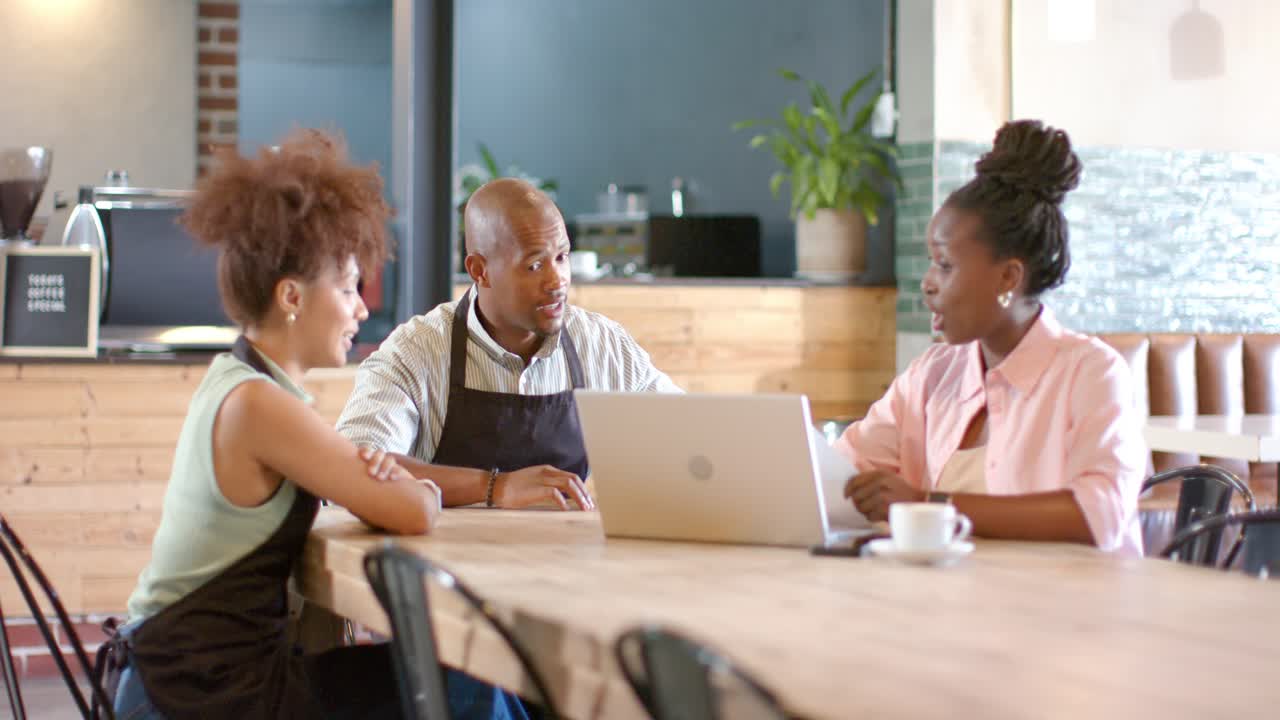 Afroamerikanische Mitarbeiter in Schürzen an einem Cafétisch, die rechte Frau öffnet ihren Laptop und plant die Schicht.