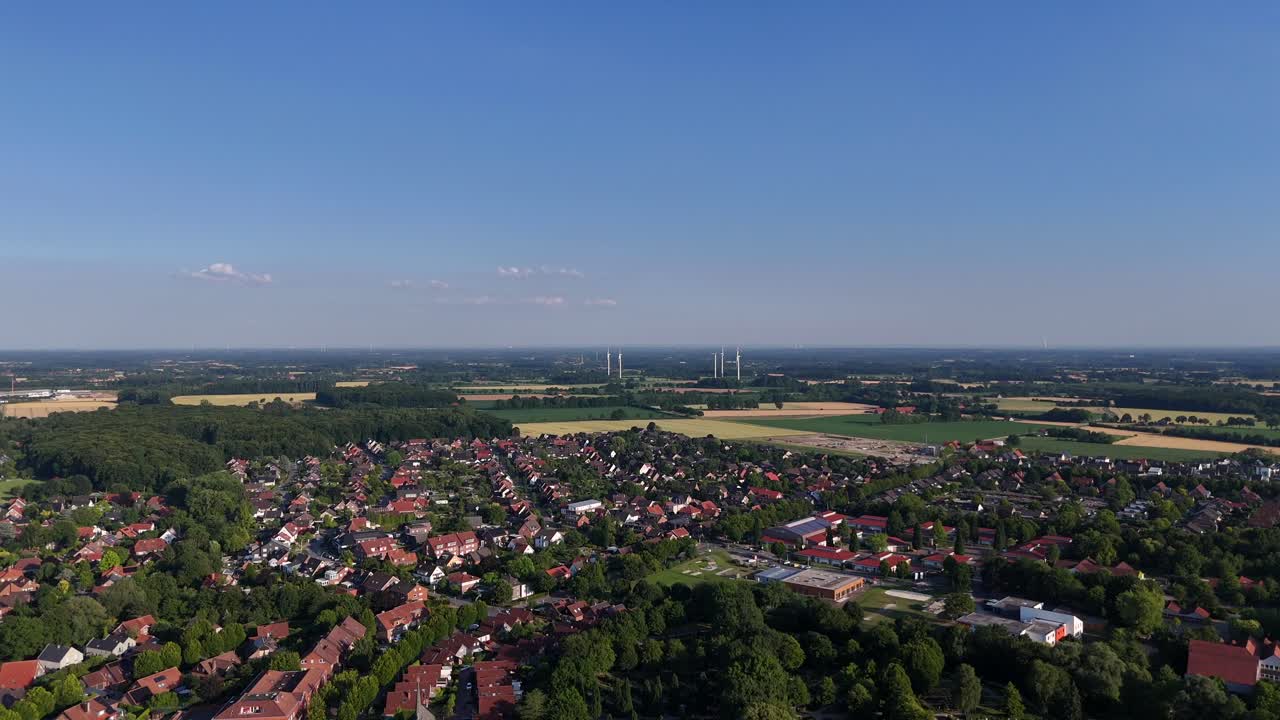 Housing area between green trees and farm fields in America. Blue sky with wind turbines in distance. Red and orange tiles on roofs of homes in small town of USA. Aerial wide shot.
