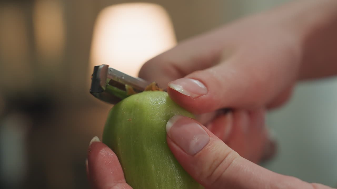 Closeup of hands peeling fresh green apple. Home cook carefully peeling green apple under warm lighting. Detailed shot of hands expertly peeling green apple in cozy kitchen setting