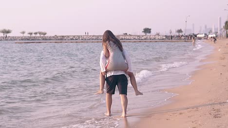 Un-Hombre-Fuerte-Lleva-A-Una-Chica-En-La-Espalda-En-Cámara-Lenta-De-La-Línea-De-Surf