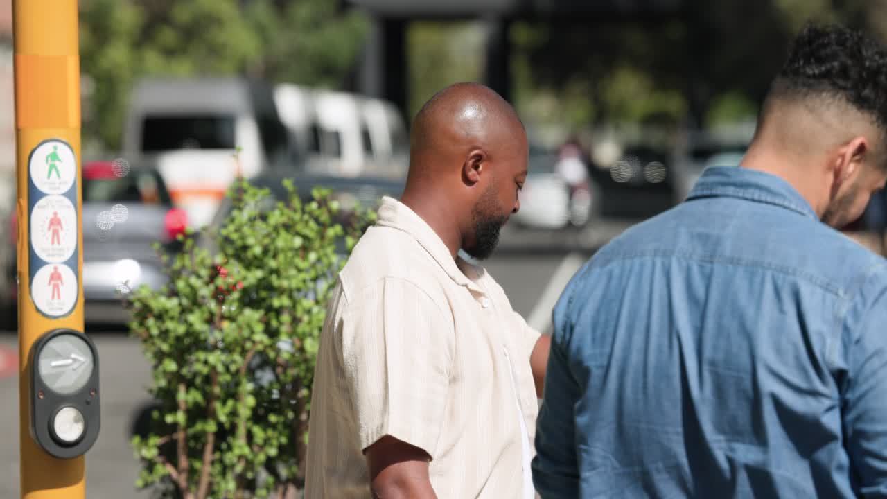 Uomini afroamericani vestiti in beige e denim sorridono al palo con pulsante per l'attraversamento pedonale, mentre camminano per attraversare la strada.