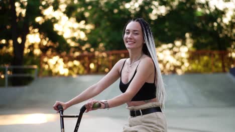 girl with black and white dreadlocks sitting on the bike in skatepark and smiling to the camera. blurred trees in sunlight on