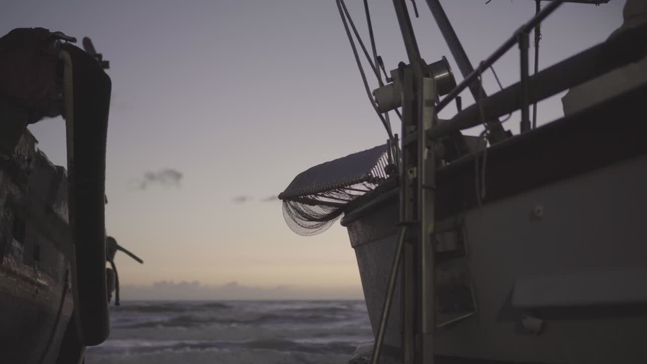 Rough sea in the blue hour from between two fishing boats parked on the shore - Trucking shot from left to right