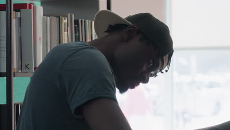 Close up side view of focused student wearing backward cap in library, leaning toward bookshelf while searching and sorting through books