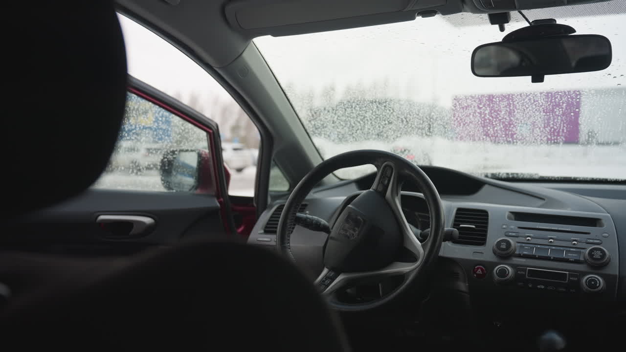 Close up of car interior showing steering wheel and wet window with water droplets, driver reflected in side mirror approaches car and opens door to enter vehicle in cold snowy urban environment