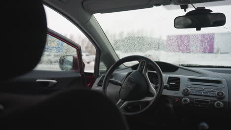 close up of car interior showing steering wheel and wet window with water droplets, driver reflected in side mirror approaches car and opens door to enter vehicle in cold snowy urban environment