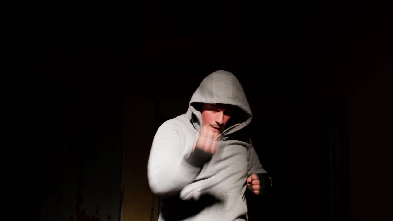 Hooded man boxing under dramatic spotlight with a black background, intense focus.