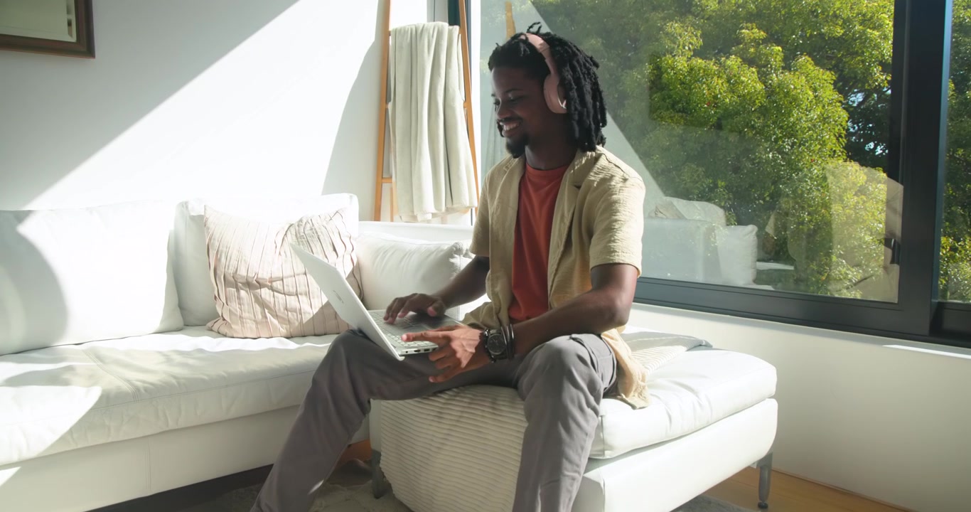 African American man putting on headphones and opening laptop, then typing on lap by window
