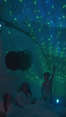 la hermana pequeña y el hermano estudian el cielo estrellado usando proyección en el techo blanco. la madre y los hijos pasan la noche acostados en una cama acogedora juntos. ocio de fin de semana