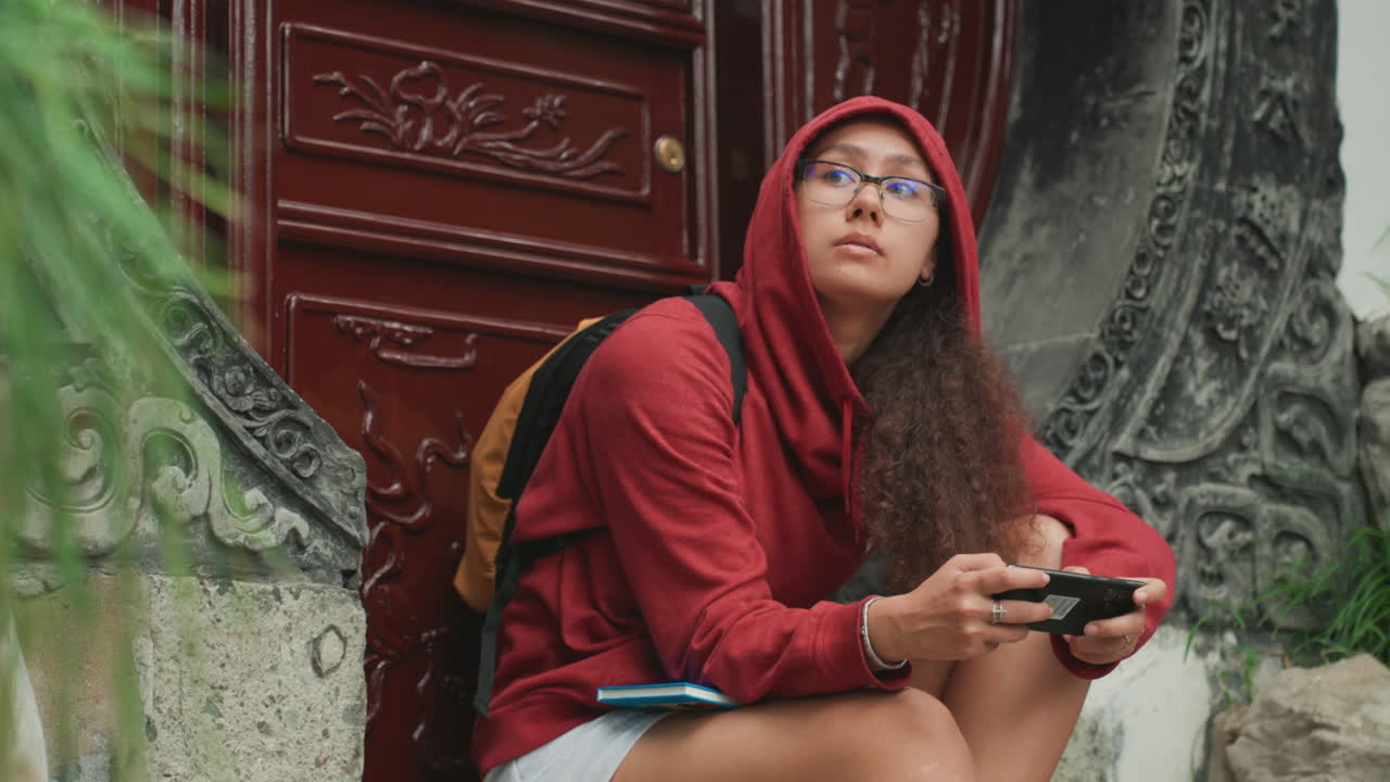 Giovane donna con felpa rossa con cappuccio studia attentamente lo schermo del telefono, occhiali che catturano la luce dello schermo, dita che scorrono con intenzione, espressione di concentrazione e curiosità, seduta accanto a un ingresso ornato