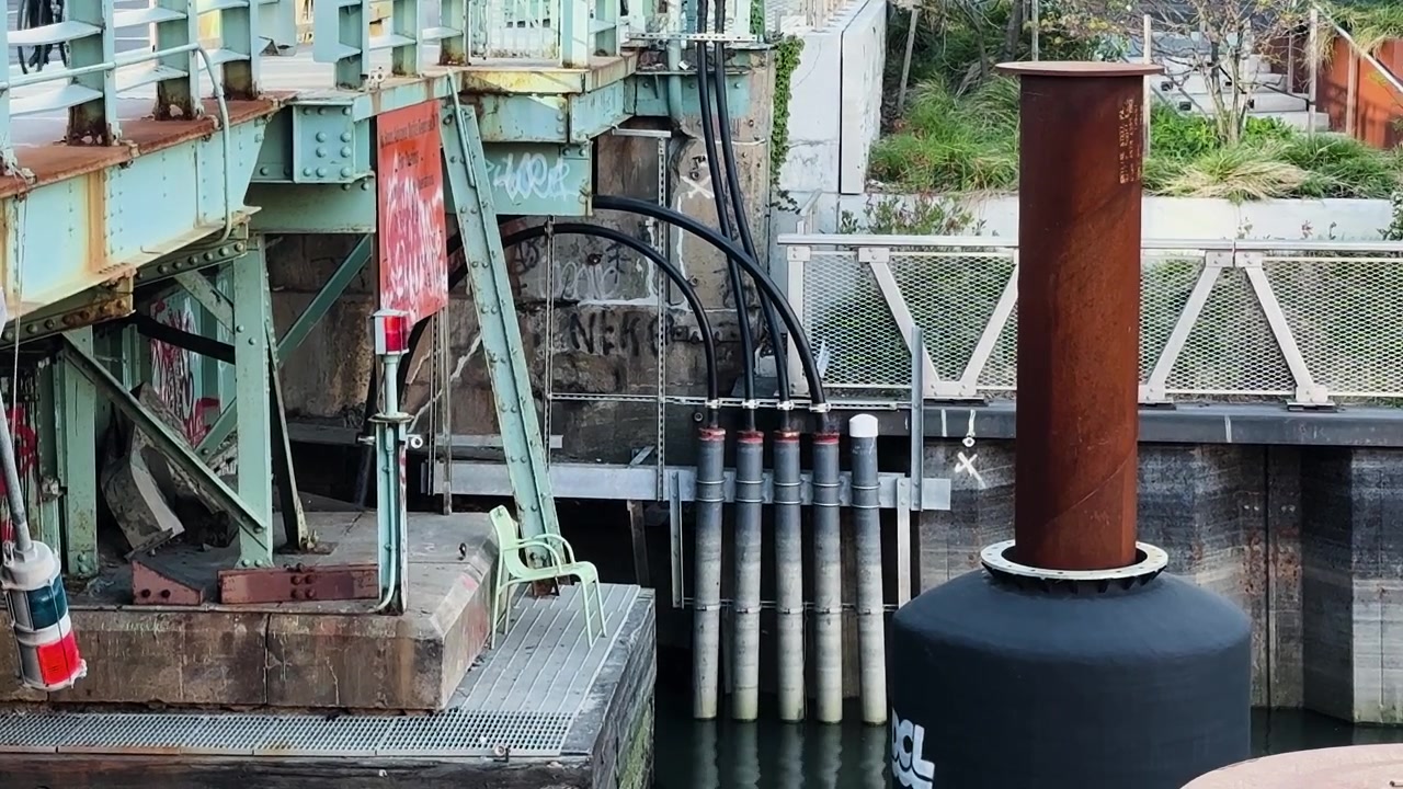 Tubi industriali e pali metallici lungo un canale sotto un ponte urbano, dettaglio di infrastruttura.