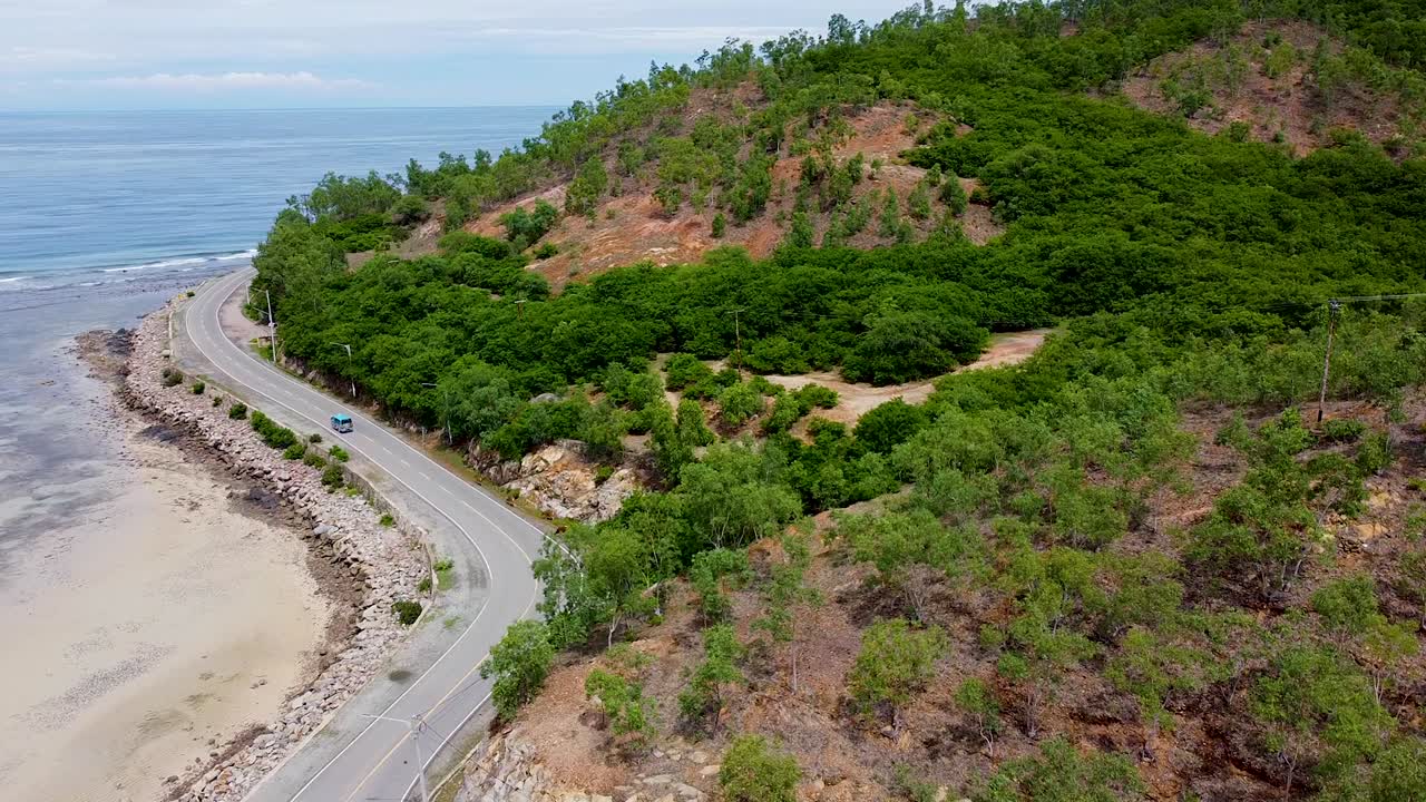 Aerial view rising over blue microlet bus traveling on coastal road next to ocean and rugged hills with trees in Dili, Timor-Leste, Southeast Asia