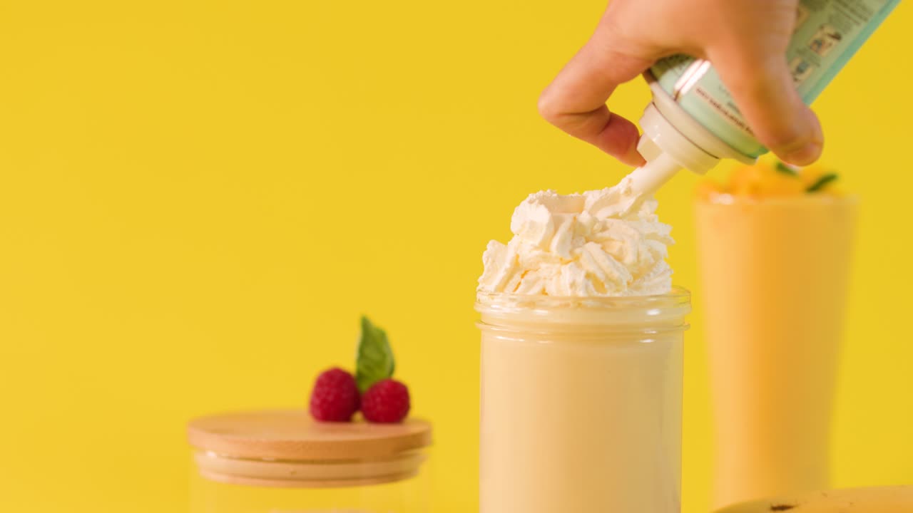 Close-up hand topping banana smoothie with whipped cream against bright yellow studio background