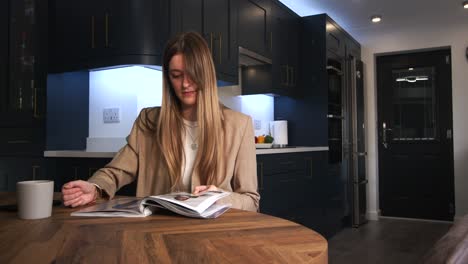 female model in a smart jacket drinking from a cup reading a magazine and looking at phone at a kitchen table in a modern kitchen