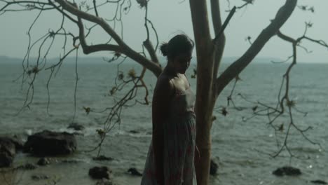 una mujer se encuentra artísticamente junto al mar bajo las ramas de un árbol, creando una silueta contra el agua