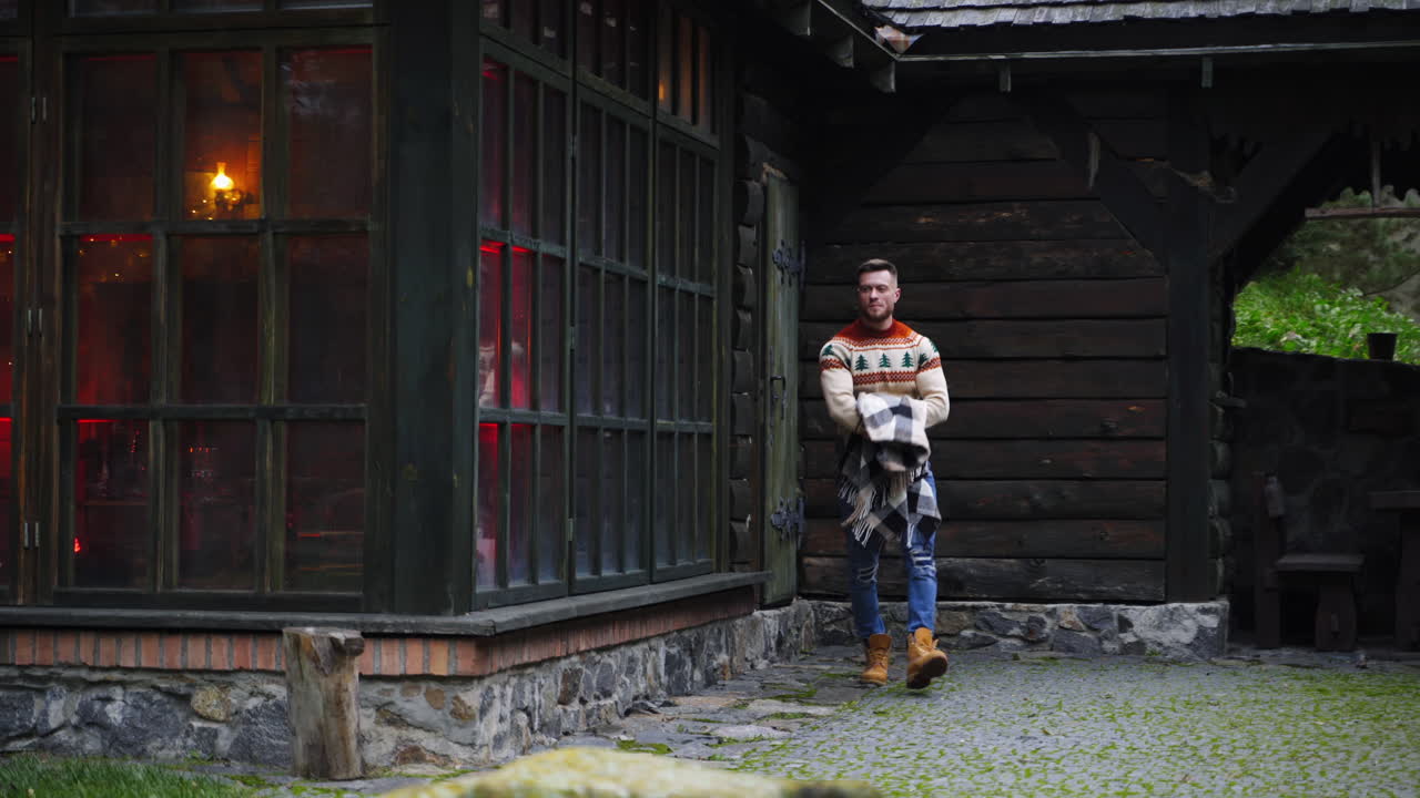 Man walks out a wooden house with big blanket in hands. Autumn evening outside.