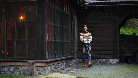 man walks out a wooden house with big blanket in hands. autumn evening outside.