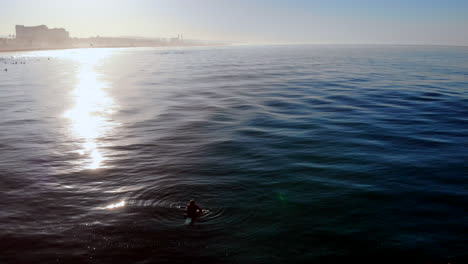 Lone-sufer-in-sunrise-silhouette-waits-for-wave-in-Pacific-Ocean-at-Huntington-Beach-California,-4k,-drone