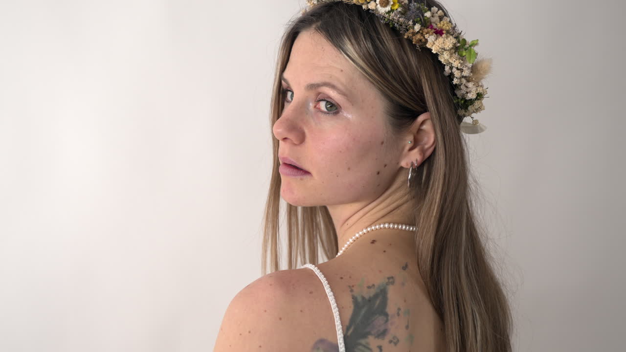 Camera tracks forwards, approaching a bride that has her back towards the lense, she has a floral crown and tattoos on her back. As the camera gets closer, she turns to face it, her expression neutral