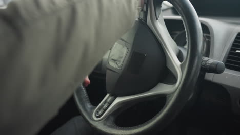 close up of person gripping car steering wheel with both hands while sitting inside parked vehicle, urban buildings slightly blurred in background under daylight through side window