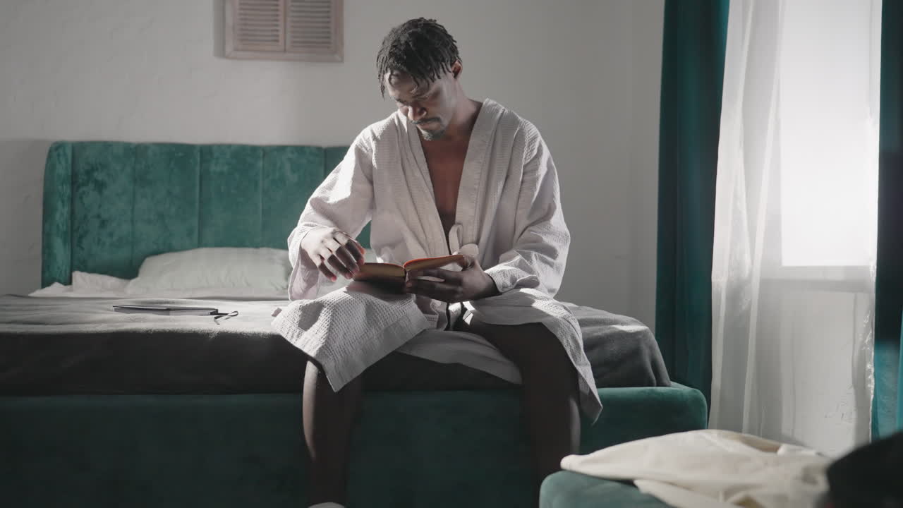 Man sitting on bed in white bathrobe, reading book with focus and concentration in well-lit room with green velvet headboard. Soft lighting creates a calm, serene atmosphere