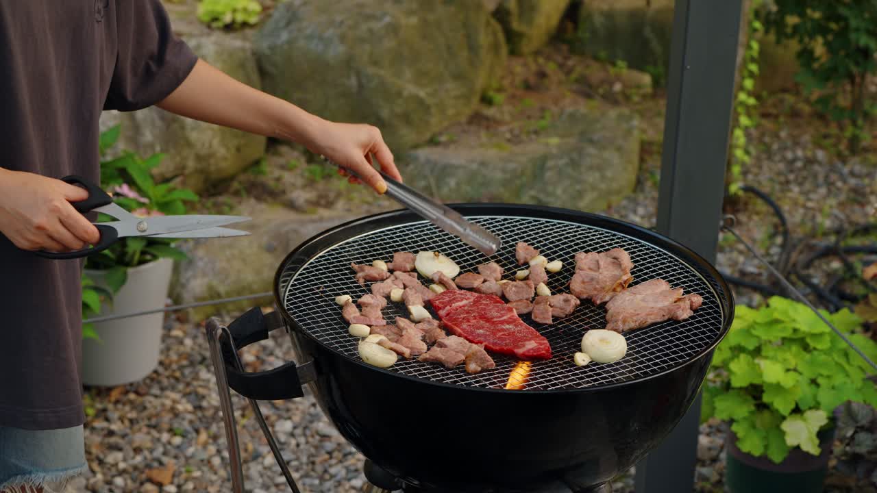 A woman uses tongs to flip and check if the bite-sized pieces of pork are cooked and ready to eat during a Korean-style barbecue in a beautiful summer garden