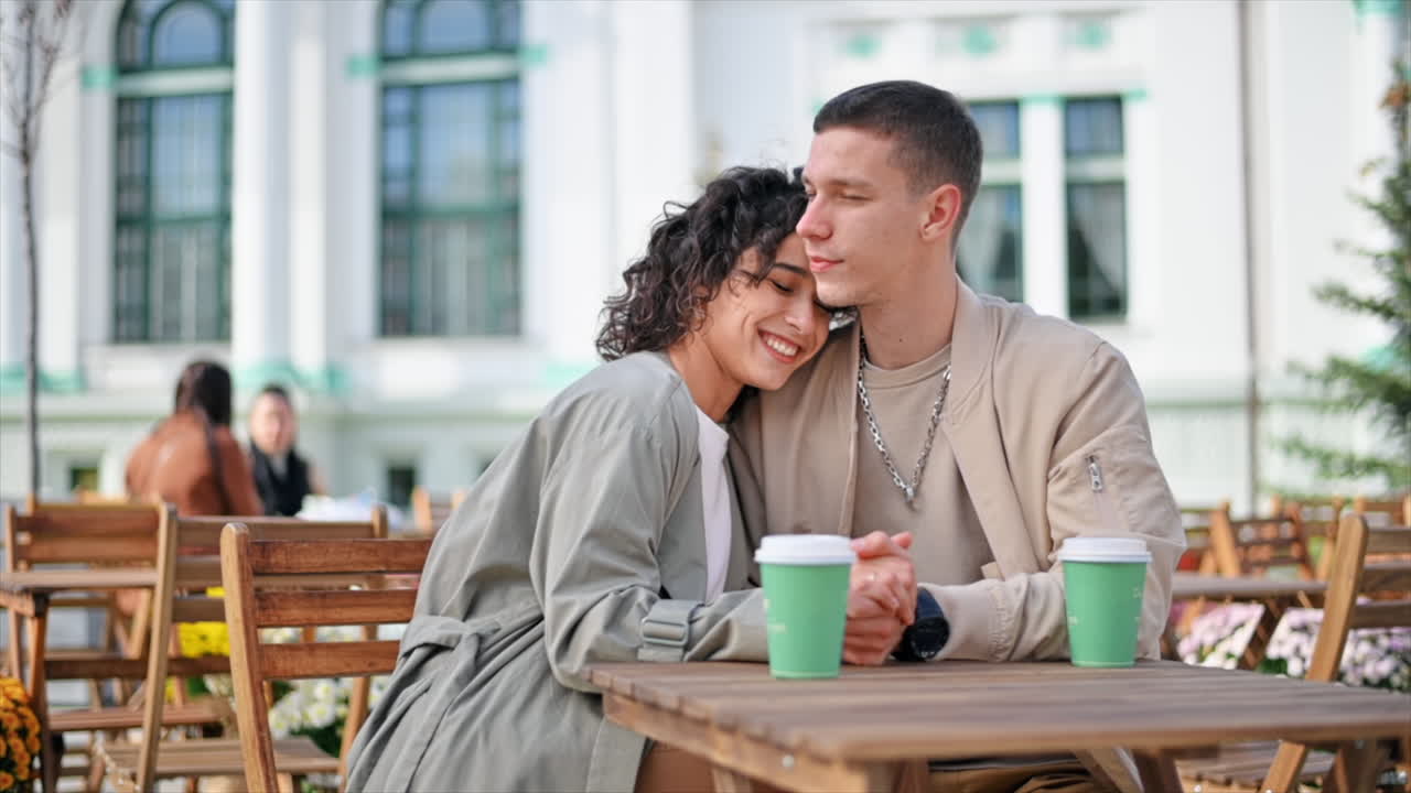 A romantic couple outdoors near a cafe. Woman lying on the man's shoulder, holding hands, smiling, coffee on the table. Autumn atmosphere. Slow motion