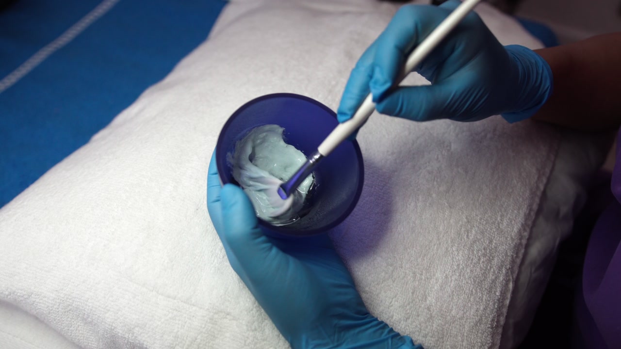 Hands of professional preparing skincare cream in bowl for spa facial treatment.