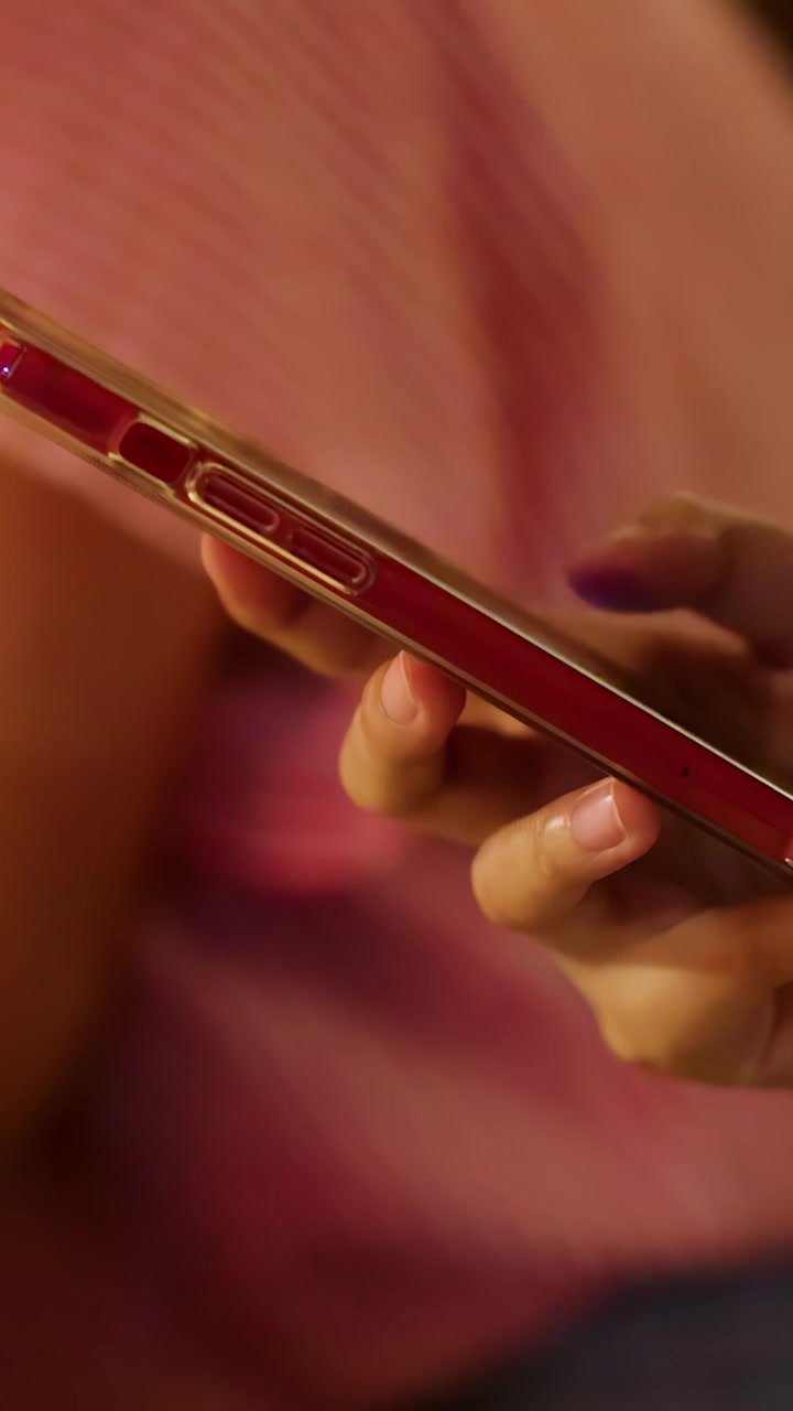 Una persona interagisce con uno smartphone rosso, tenendo e toccando il dispositivo in un ambiente interno illuminato caldamente, indossando una camicia a righe rosa