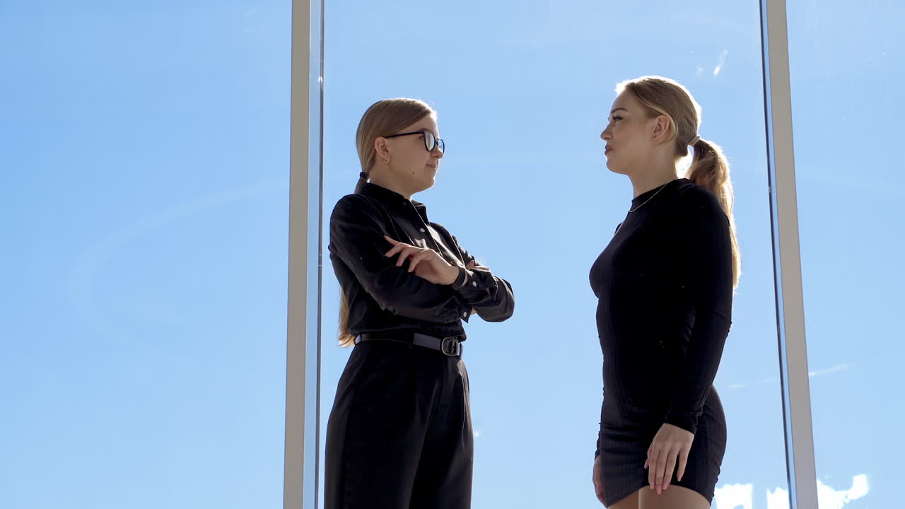 Beautiful girls talking together near the window. Pleasant conversation between two young woman wearing black on clear sky background indoors. Profile view.