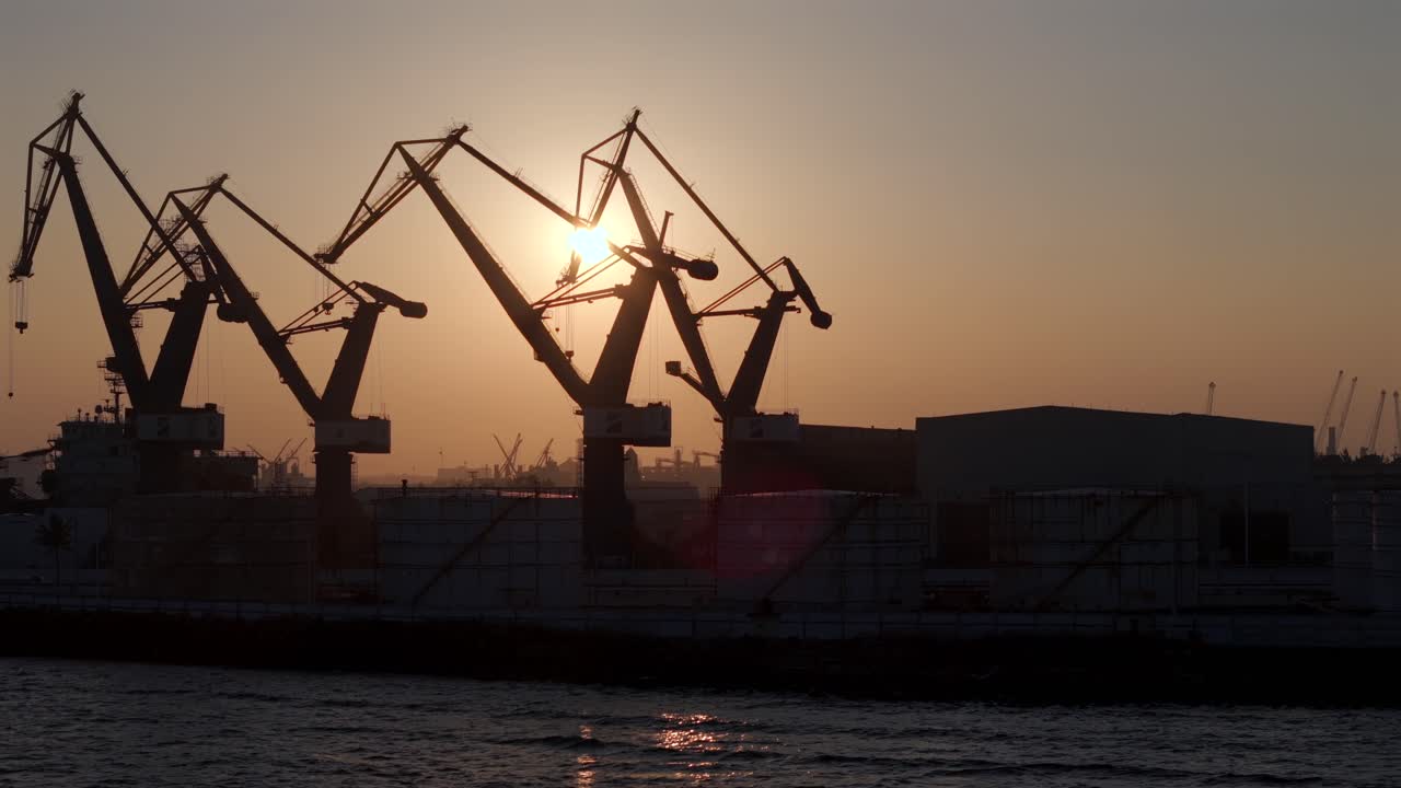 Rotating shot of shipyard cranes in silhouette against the setting sun. Strong backlight highlights their structure against the sky.
