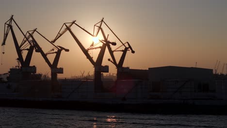 rotating shot of shipyard cranes in silhouette against the setting sun. strong backlight highlights their structure against the sky.