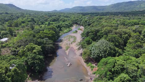 Aerial-drone-shot-over-the-Guanuma-River-in-Yamasá,-Monte-Plata:-lush-green-areas,-majestic-mountains,-blue-sky,-and-gentle-clouds-on-a-sunny-day,-capturing-the-natural-beauty-of-the-region