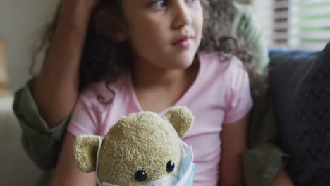 madre e hija de raza mixta abrazándose, hija sosteniendo un oso con máscara facial